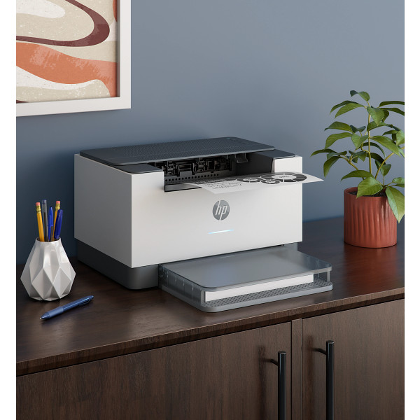 HP LaserJet M211dw Light Basalt Product sitting on desk aganst blue wall, right facing, vertical ori